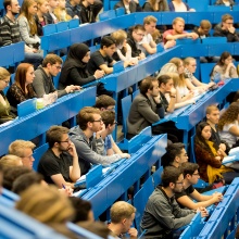 Students sit in the lecture hall on blue lecture hall benches. 