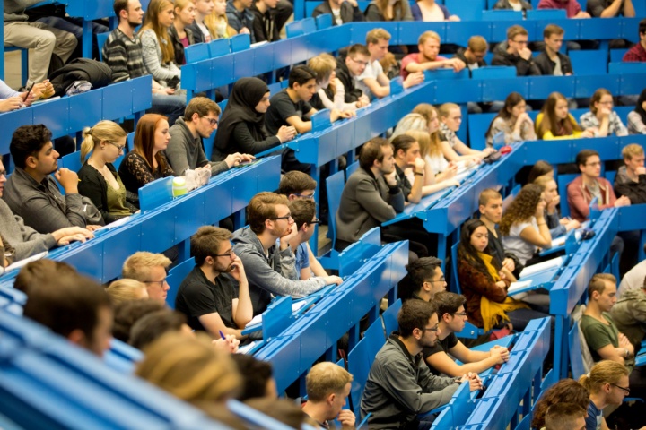 Students sit on the blue benches of a lecture hall and look forward attentively.